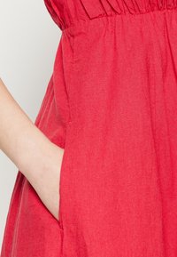 Robe en lin rouge avec taille froncée et poches latérales. Le tissu a une surface texturée avec une douce drapé.