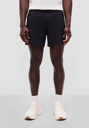 Mand iført sorte sportsshorts, hvide sokker og hvide sneakers, står opret mod en ensfarvet grå baggrund.