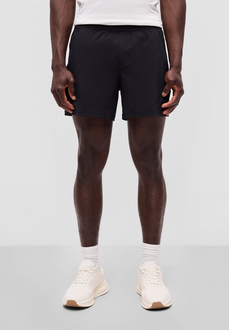 Uomo che indossa pantaloncini sportivi neri, calzini bianchi e scarpe da ginnastica bianche, in piedi dritto contro uno sfondo grigio uniforme.