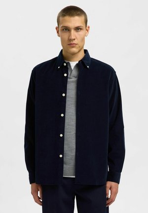 Marineblauwe corduroy knoopshirt met een klassieke kraag, voorzien van witte knopen, gelaagd over een lichtgrijs sweater. Soepele textuur, relaxte pasvorm.