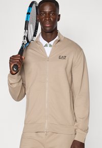 Bēša rāvējslēdzēja jaka no mīksta auduma ar stāvu apkakli un logotipa izšuvumu krūtīs, turot tenisa nūju.