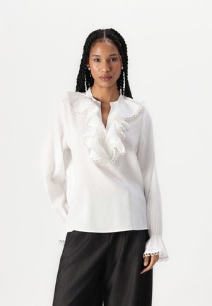 Blouse blanche avec col volanté et devant, présentée avec un bord en dentelle. Manches longues avec poignets froncés. Portée avec un pantalon large et sombre.