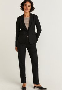 Musta räätälöity bleiseri kahdella etupussilla, yhdistettynä mustiin slim-fit-housuihin. Alla näkyy vaalea beige paita. Tyylikäs, rakenteellinen muotoilu.