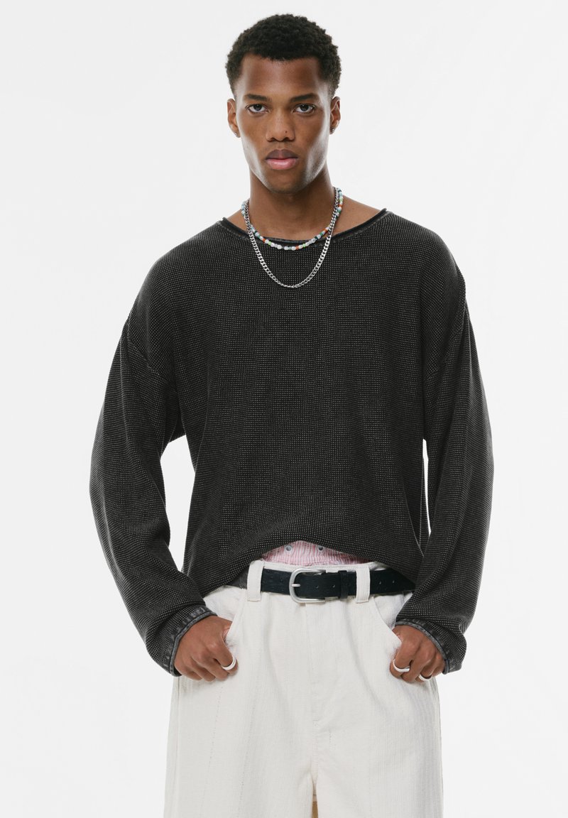 Zwarte longsleeve top van een gestructureerde stof, ontspannen pasvorm, brede halslijn; gedragen met witte broeken en gelaagde kettingen voor contrast.