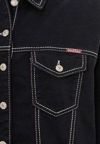 Schwarze Jeansjacke mit kontrastierendem weißen Stich, Metallknöpfen und einem roten Etikett an der Tasche. Zwei Vordertaschen mit Klappen.