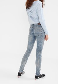 Sudadera azul claro, jeans ajustados de lavado claro con una sutil textura desvanecida, y zapatillas negras con suelas blancas. Vista desde la parte de atrás.