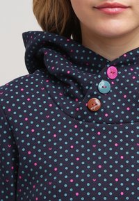 Marinblått tyg med prickar i rosa och turkos. Har en bred krage och tre knappar i rosa, ljusblått och brunt.