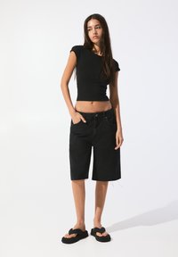 Zwarte fitted t-shirt, licht-in-waisted zwarte denim shorts en zwarte sandalen. Shorts hebben een rafelige rand; het model staat met één hand in de zak.