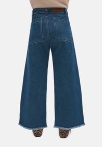 Vaqueros de mezclilla azul de pierna ancha con bajo deshilachado, que cuentan con una cintura alta, dos bolsillos traseros y una etiqueta de cuero marrón en la cinturilla.