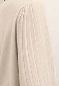 Helles beiges Sweatshirt mit gerippten Falten an den Ärmeln, das eine weiche Textur und eine figurbetonte Passform aufweist. Nahaufnahme der Ärmeldetails.