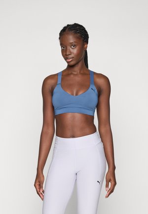 Blue sports bra with thick straps, soft fabric, and a rounded neckline. Paired with light gray high-waisted leggings featuring a logo.