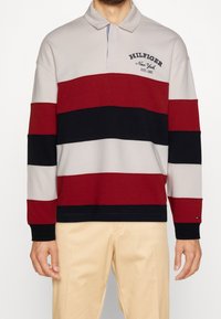 Polo a maniche lunghe a righe nei colori rosso, blu navy e grigio; presenta un colletto e un logo ricamato sul petto. Tessuto morbido, vestibilità comoda.