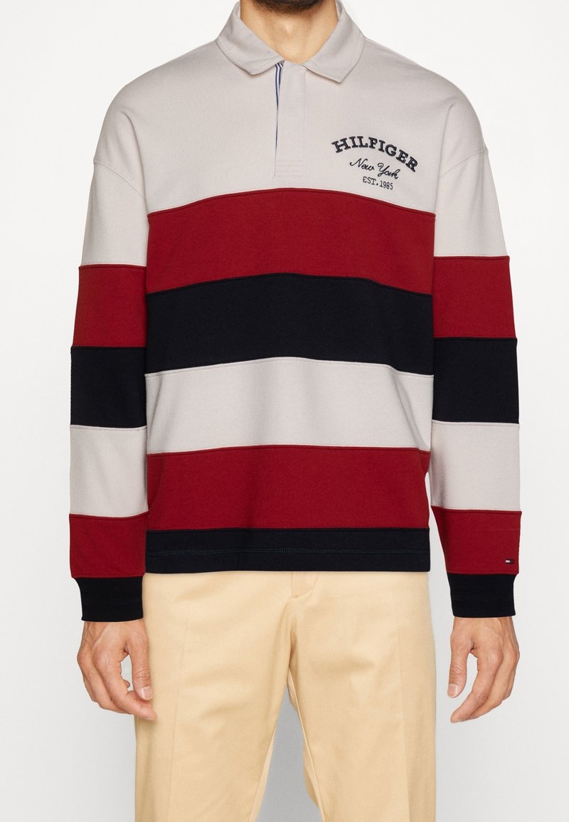 Polo a maniche lunghe a righe nei colori rosso, blu navy e grigio; presenta un colletto e un logo ricamato sul petto. Tessuto morbido, vestibilità comoda.