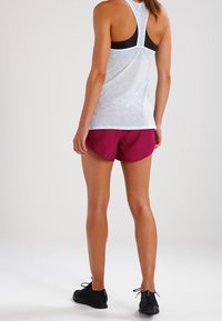 Světle šedé atletické tílko bez rukávů s černými detaily, kombinované s tmavě červenými šortkami a černými atletickými botami, pohled zezadu.