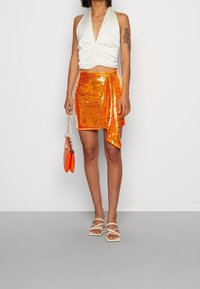 Top blanco sin mangas con un frente fruncido, combinado con una falda corta de lentejuelas naranjas brillantes. Sandalias beige con tiras y un pequeño bolso naranja.