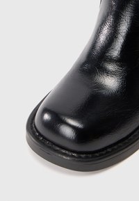 Bota de cuero negro con acabado brillante, puntera redondeada y una suela de goma negra resistente, con costuras mínimas a lo largo del borde.