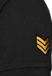 Schwarzer Strickstoff mit strukturierter Oberfläche, versehen mit einem goldenen bestickten Chevron-Emblem auf dem linken Ärmel.