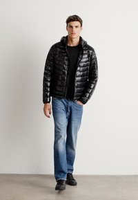 Zwarte pufferjas met een glanzende textuur, capuchon en ritssluiting, gedragen over een zwart shirt, gecombineerd met lichtblauwe spijkerbroek en zwarte schoenen.