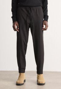 Pantaloni jogger negri, cu șnur la brâu, manșete elastice și textură moale, asortați cu cizme bej până la gleznă pe un podea neutră.