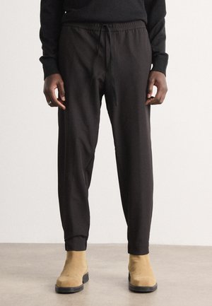 Black, drawstring jogger pants with elastic cuffs and soft texture, paired with tan ankle boots on a neutral floor.