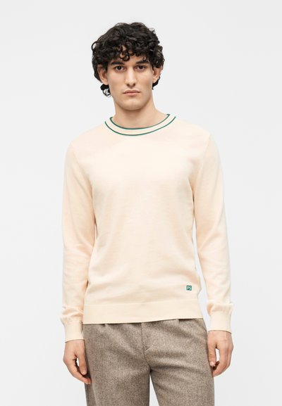 Pull long en pêche clair avec un col rond, bordé de vert et un petit logo bleu en bas à gauche. Texture douce.