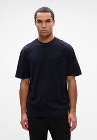 Μαύρο κοντομάνικο μπλουζάκι από βαμβάκι με στρογγυλή λαιμόκοψη, διαθέτοντας τσέπη στο στήθος και λεπτομέρεια ετικέτας στο πλάι σε πορτοκαλί χρώμα.