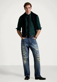 Άνδρας με πράσινο καρό πουκάμισο, μαύρη γραβάτα, αμάνικο πουλόβερ κρεμασμένο στους ώμους, σκισμένα μπλε τζιν, μαύρες αθλητικές υποδημασίες, χέρια στις τσέπες.