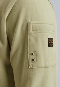 Felpa verde in cotone con tasca sul petto con pattina, due occhielli metallici e un'etichetta in tessuto rettangolare cucita sul lato.