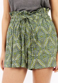 Shorts verts à motifs avec une ceinture volantée et un nœud à l'avant, ornés d'un design floral et géométrique avec des accents jaunes et blancs.