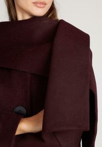 Bordeaux wollen jas met een hoge kraag, grote schouderflap, grote zwarte knoopdetails en een zachte, gestructureerde stof.