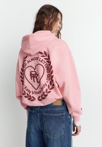 Felpa rosa con un grande motivo marrone che raffigura un cuore e del testo sul retro, abbinata a jeans blu a vita alta.