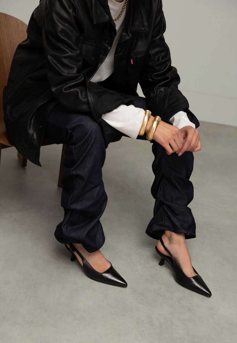 Personne assise sur une chaise en bois portant un jean bleu foncé, une veste en cuir noire, une chemise blanche, des bracelets en or et des escarpins noirs à bout pointu.