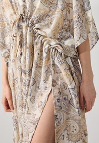 Robe longue imprimée en tissu léger et fluide avec un motif paisley en gris et beige. Elle présente des détails froncés et une fente sur le côté.