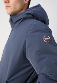Mornarsko modra vodoodporna jakna s kapuco. Značilna gladka tekstura in okrogla Colmar nalepka na levi rokavu.