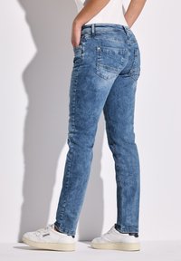 Persona que lleva jeans azul desgastados y zapatillas blancas, de pie contra un fondo blanco con las manos en los bolsillos.