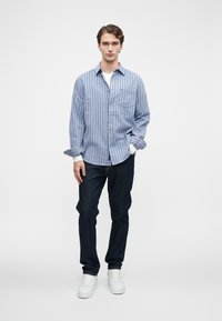 Blauw-wit gestreepte langesleeve shirt, met knopen en een borstzak. Draagt met donkere jeans en witte sneakers, op een effen achtergrond.