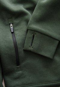Mørkegrøn sweatshirt stof har en lynlåslomme og en ribbet manchet. Materialet ser blødt ud med en glat tekstur.