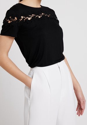 Kobieta w czarnej bluzce z krótkim rękawem ozdobioną koronką na piersi i rękawach, w połączeniu z białymi spodniami z wysokim stanem, z ręką w kieszeni.