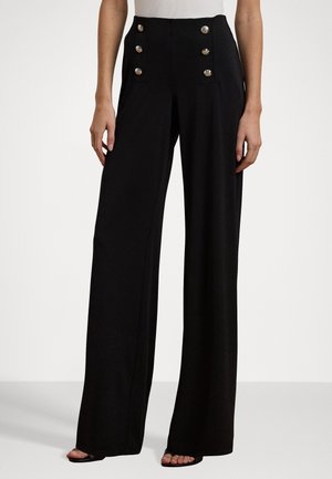 Schwarz weite Hose mit hohem Bund und sechs metallischen Knöpfen vorne, getragen mit weißem Oberteil und schwarzen Sandalen mit offenen Zehen.