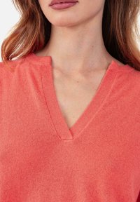 Pull col en V corail en tissu tricoté doux. Présente une texture côtelée subtile et un ourlet non fini au niveau du col pour un look décontracté.