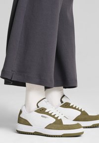 Baskets blanches et vert olive avec une tige en suède et cuir, dotées de lacets blancs et d'une semelle en caoutchouc texturée, assorties à un pantalon gris côtelé.