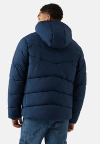 Granatowa kurtka puchowa z kapturem, o pikowanej fakturze, poziomym przeszyciem oraz naszywką z logo na lewym ramieniu.