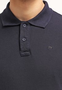 Marinblå polo-shirt med en texturerad tyg, tvåknappsknäppning och en liten logotyp på vänster sida av bröstet.