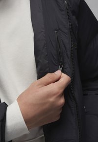 Sort pufferjakke med en lynlås-lomme, glat tekstur og opretstående krave. En hvid sweatshirt er synlig under jakken.