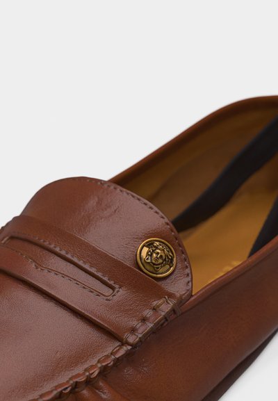 Mocassin en cuir marron avec bride décorative et emblème circulaire doré présentant une tête de Méduse sur le dessus avant.