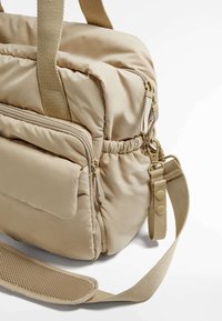 Borsa a mano imbottita beige con manici superiori doppi, tracolla regolabile, tasca frontale con zip e tessuto leggermente testurizzato. Chiusura con zip e dettagli hardware laterali.