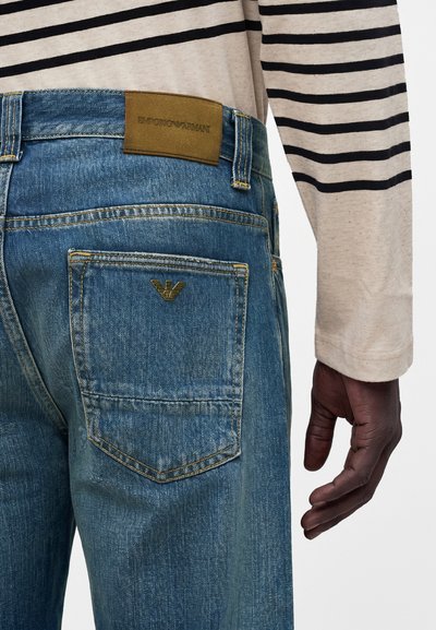 Άτομο που φοράει μπλε τζιν denim με καφέ δερμάτινη ετικέτα Emporio Armani και λογότυπο στην πίσω τσέπη, και μπεζ μακρυμάνικη μπλούζα με μαύρες ρίγες.