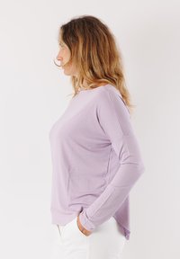 Camicia a maniche lunghe viola chiaro con scollo rotondo, texture morbida, vestibilità ampia e fessure laterali, indossata con pantaloni bianchi.