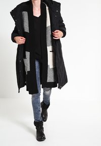 Parka nera con cappuccio, dotata di una fodera interna a fantasia, indossata sopra un top nero e jeans multicolore, abbinati a stivali neri alla caviglia.
