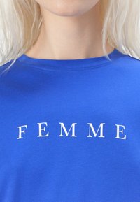 Blauwe katoenen t-shirt met een ronde hals en het woord "FEMME" in witte hoofdletters op de borst gedrukt. Gladde textuur.